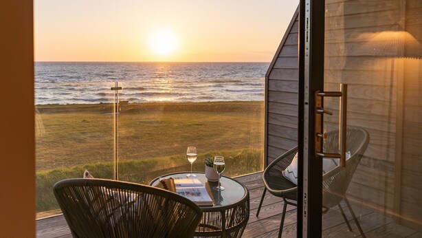 A balcony with a table set for two, overlooking the ocean at sunset. - The Lighthouse is an end of terrace four bedroomed seafront retreat with a hot tub and incredible vi (Widemouth Bay)