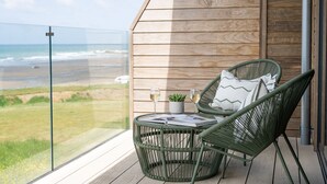A balcony with a view of the beach, a wicker chair, a small table with a plant, and a glass of wine. - The Lighthouse is an end of terrace four bedroomed seafront retreat with a hot tub and incredible vi (Widemouth Bay)