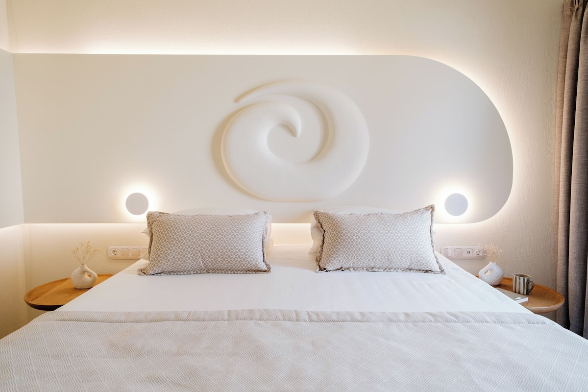 A modern hotel room with a large bed, two bedside tables, a wall-mounted light fixture, and a decorative wall piece.