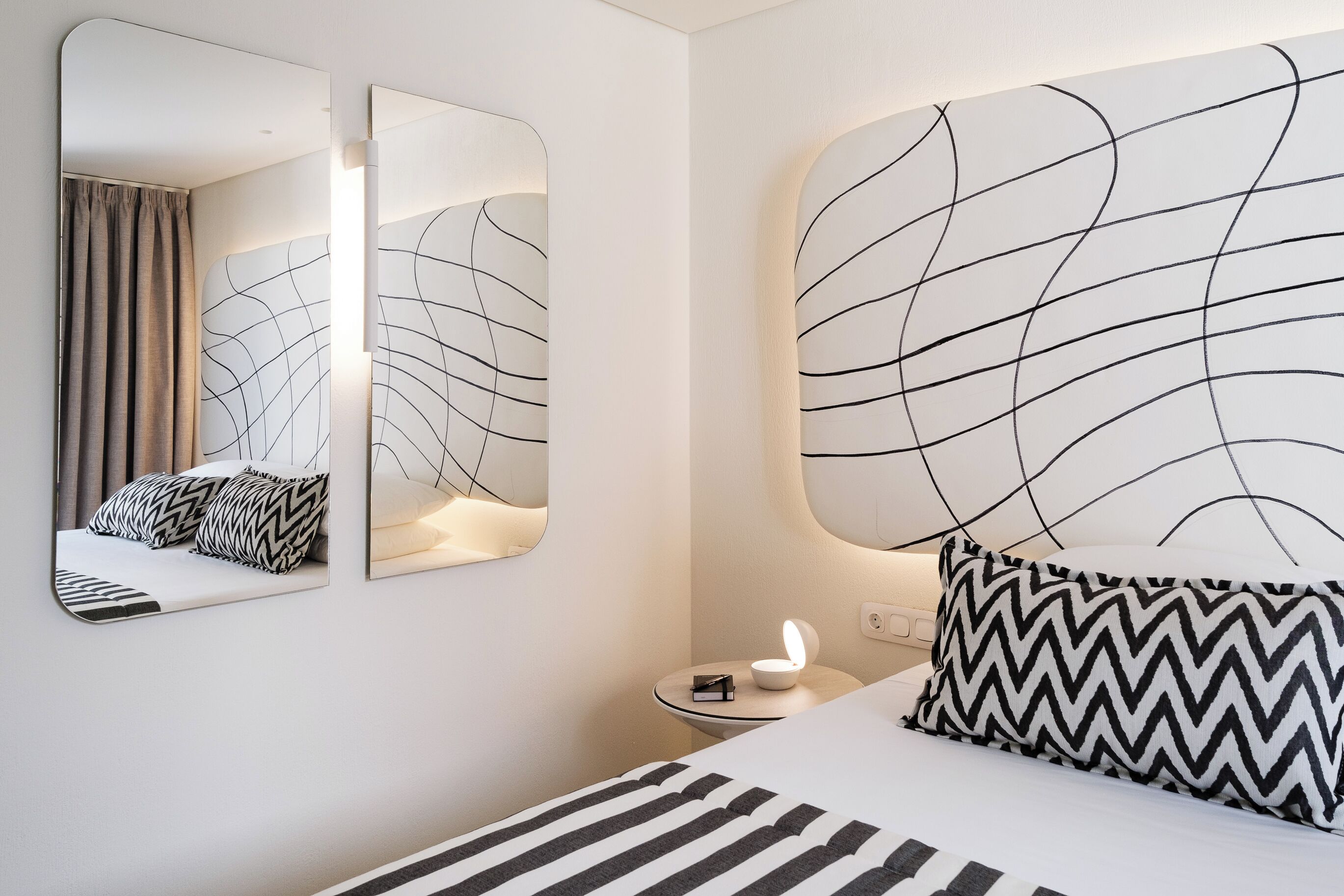 Quarto de hotel moderno com uma cama grande, uma mesa de cabeceira com abajur, um espelho e uma parede com arte abstrata em linhas.