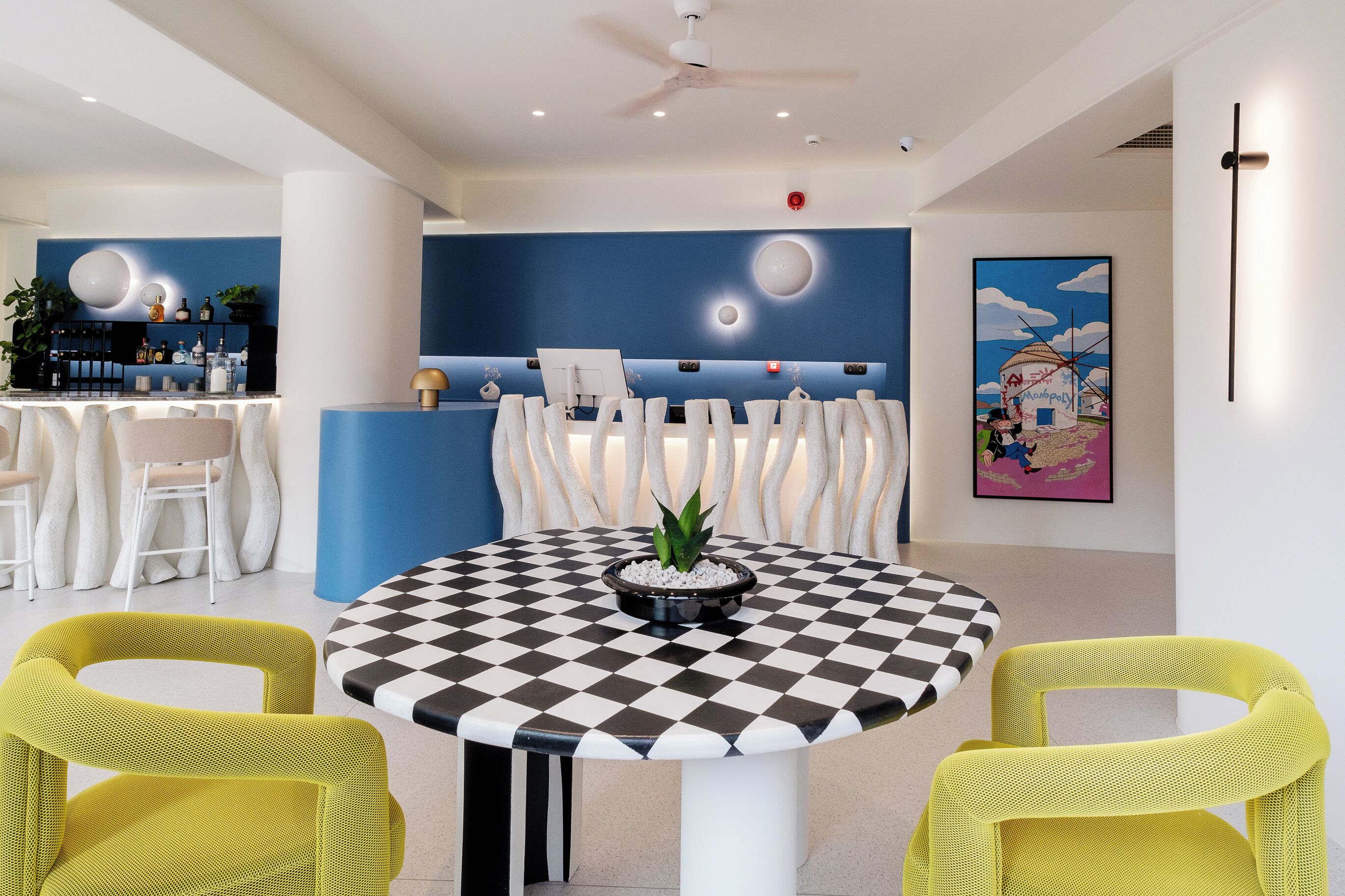 A modern lobby with a checkered table, yellow chairs, a reception desk, and a painting on the wall.