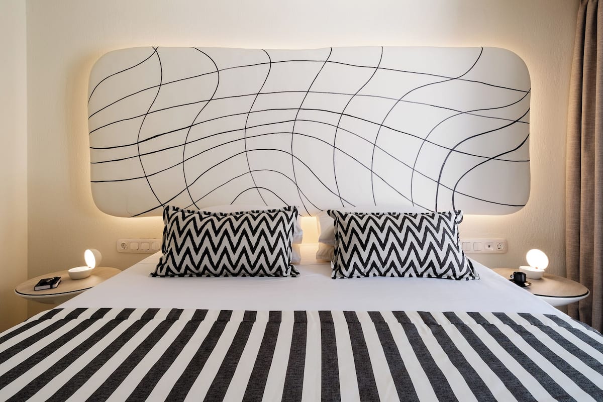A hotel room with a large bed, two bedside tables, a modern headboard, and a nightstand with a lamp.