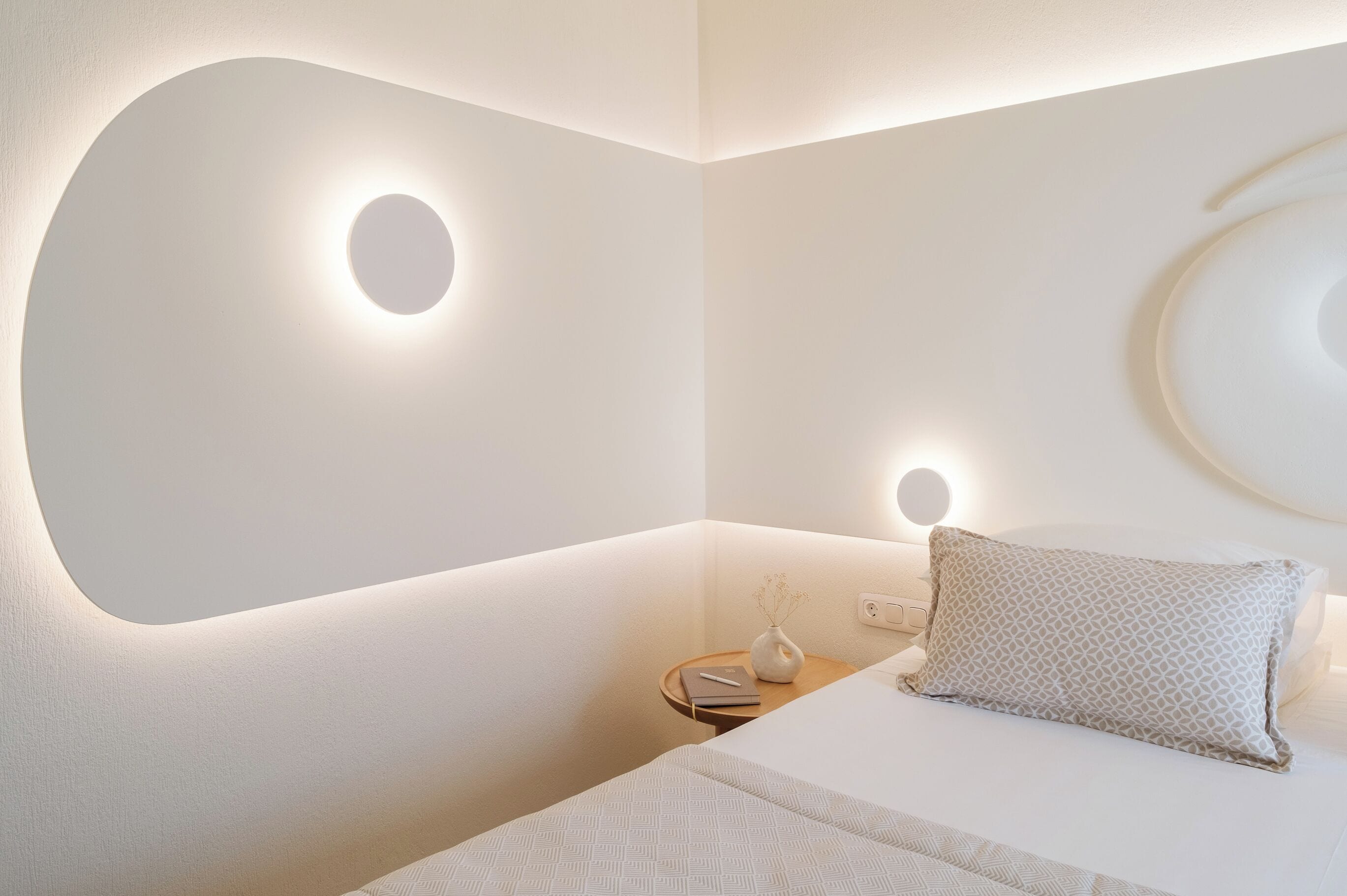 Une chambre à coucher moderne avec un mur aux lignes courbes, une table de chevet avec une lampe et un lit bien fait, agrémenté d’un oreiller.