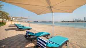 A beachside lounge area with sun loungers and umbrellas, a paved walkway, and a view of the city skyline across the water. - Pool View 1BR on Palm Jumeirah (Dubai)