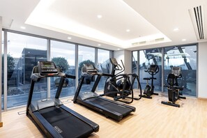 A modern gym with treadmills, elliptical machines, and a large window offering a city view. - Stunning Corner Suite Canal View (Dubai)