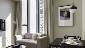 Een moderne hotelkamer met een bank, een eettafel voor twee personen en een groot raam met gordijnen.