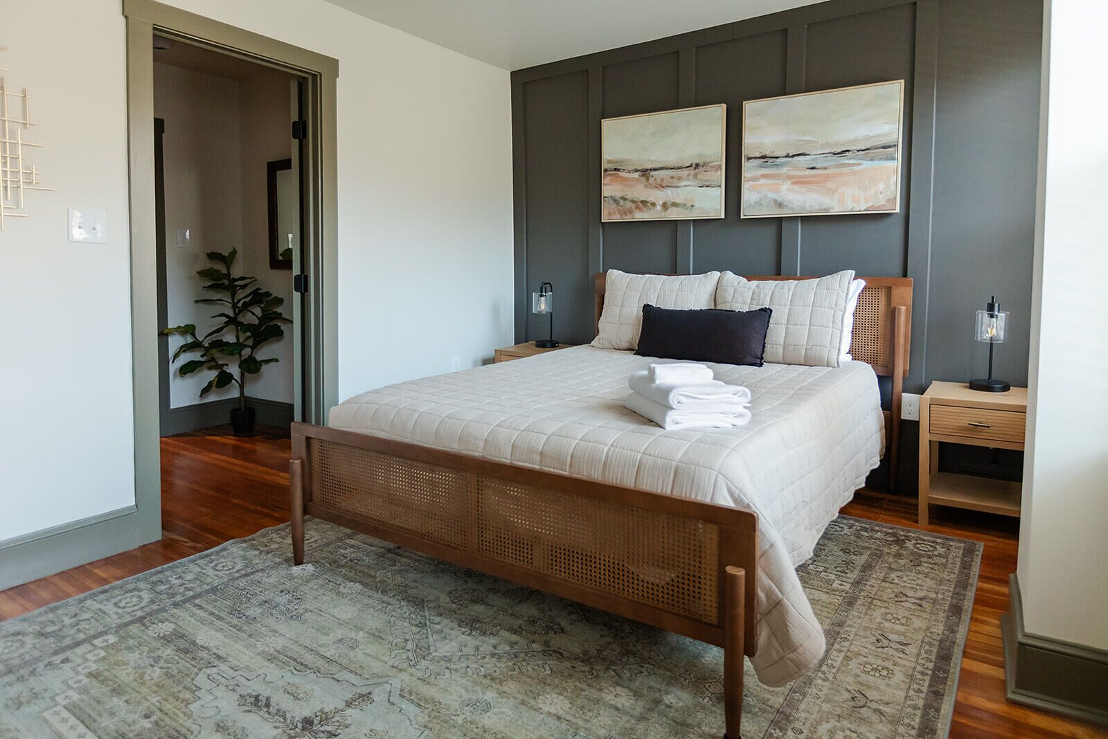 A bedroom with a large bed, a bedside table, a nightstand, a wall-mounted lamp, and a potted plant.