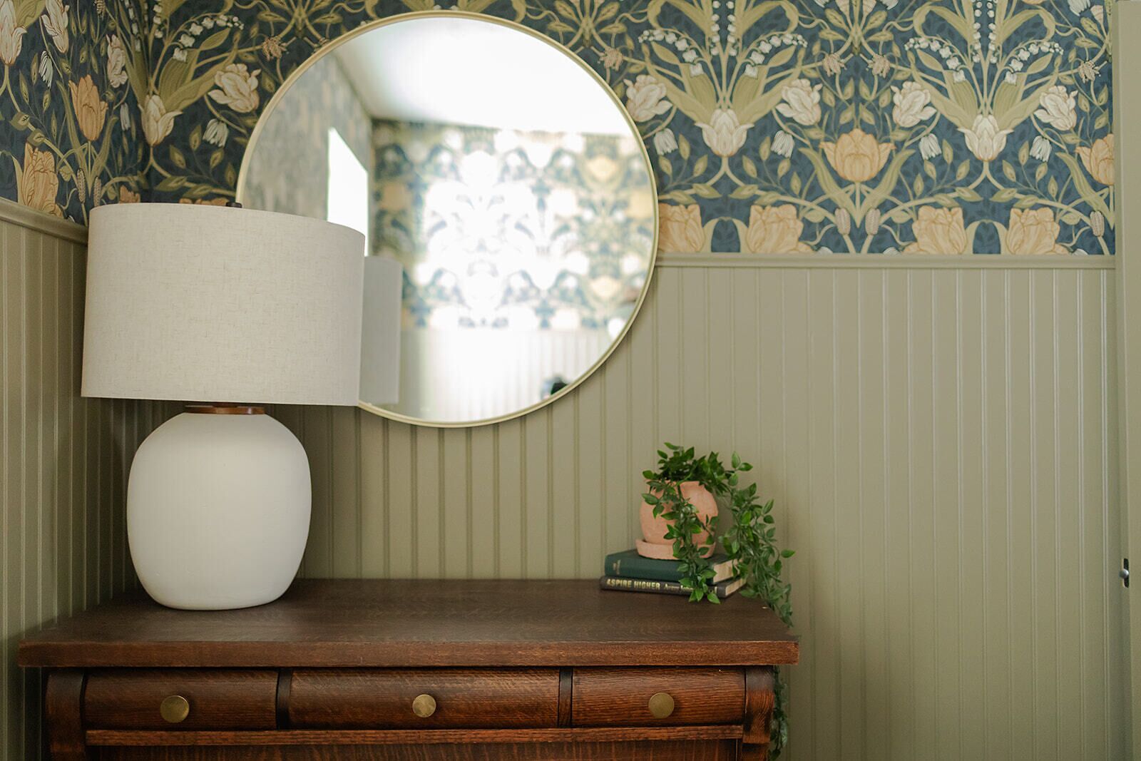Une commode en bois, surmontée d’un miroir rond et accompagnée d’une lampe blanche.
