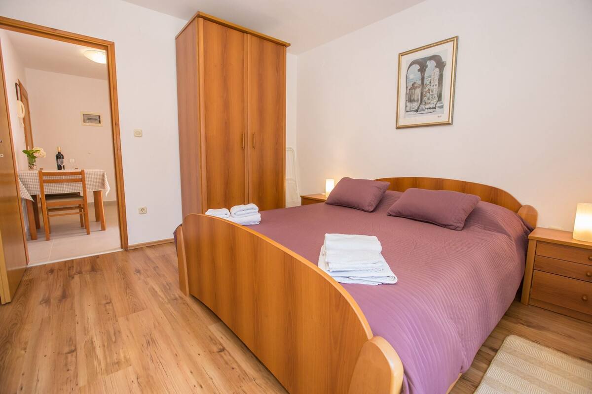 Ein Schlafzimmer mit einem hölzernen Bett, einem Nachttisch, einem Schrank, einem Spiegel, einem gerahmten Bild an der Wand und einem Essbereich mit Tisch und Stühlen.