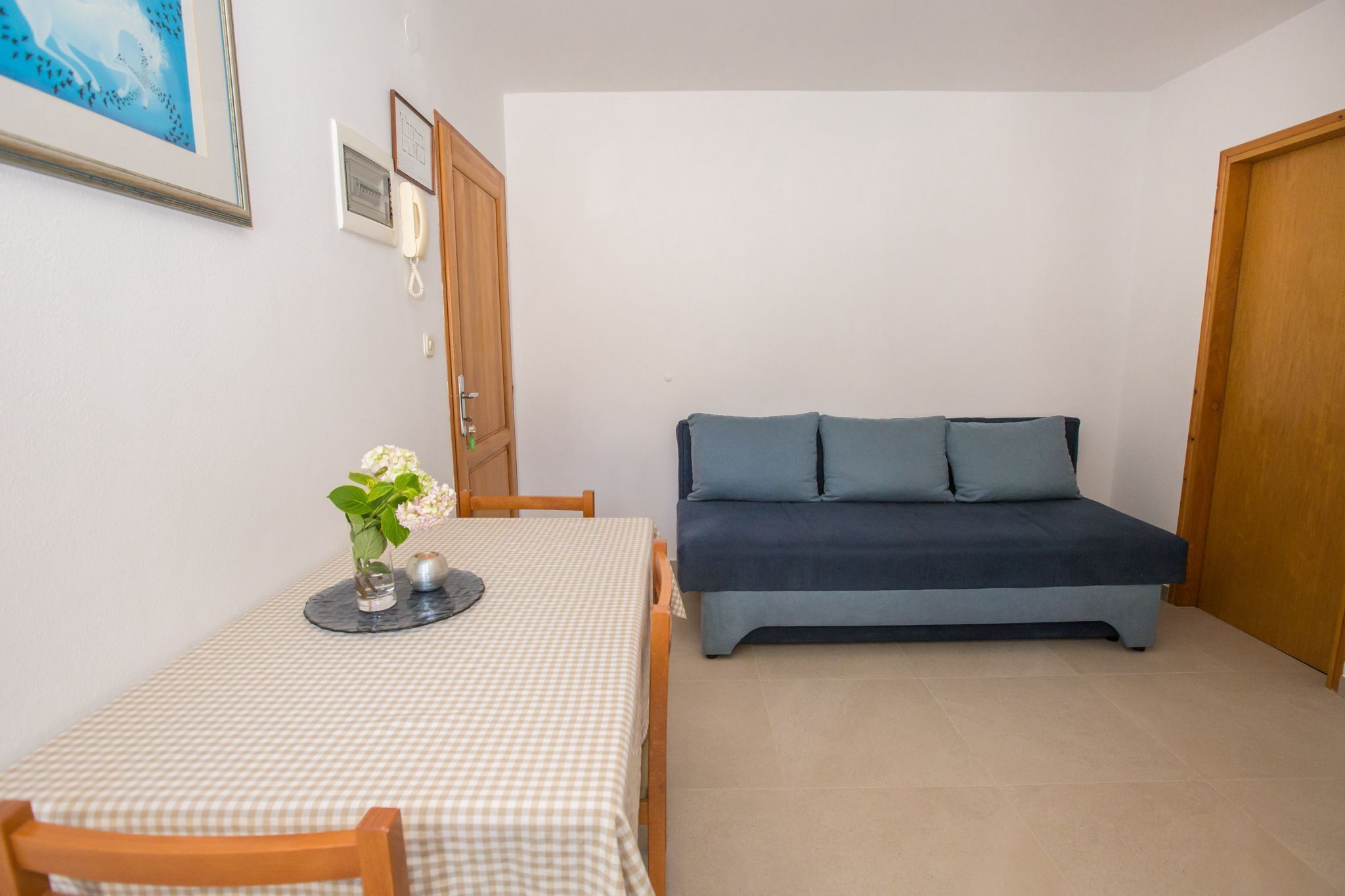 Ein kleiner, ordentlicher Raum mit einem Bett, einem Tisch mit kariertem Tischläufer, einer Blumenvase und einem gerahmten Bild an der Wand.