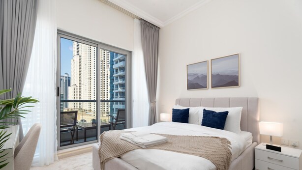 Hotelkamer met een groot bed, nachtkastje, stoel, balkon met uitzicht op hoge gebouwen, en twee ingelijste foto's aan de muur.