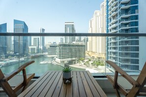 Un balcon doté de meubles en bois et agrémenté d’une plante en pot, offrant une vue sur un paysage urbain avec de hauts immeubles et un port de plaisance.