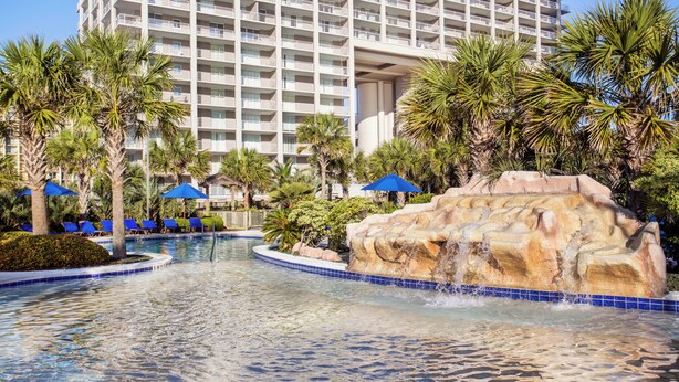 A hotel pool with a rock waterfall, palm trees, and blue umbrellas. - Ocean Front, 1602 Royale Palms, full resort access!! (Myrtle Beach)