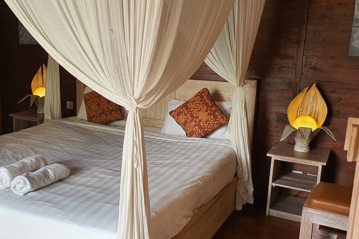 Een bed met witte lakens en een hemelbed, een nachtkastje met een lamp en een houten nachtkastje met een vaas.
