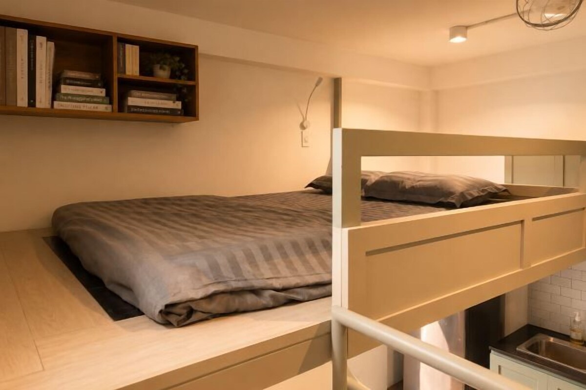 Una cama individual con estructura de madera, una mesita de noche, un estante con libros y una lámpara de pared.