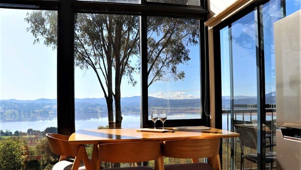 A dining area with a table and chairs, large windows with a view of a lake and mountains, and a modern interior design. - The Tamar Ridge Lookout at Tamar Ridge Apartments (Grindelwald)