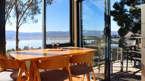 A modern dining area with a wooden table and chairs, large windows with a view of a lake and mountains, and a deck with outdoor seating. - The Tamar Ridge Lookout at Tamar Ridge Apartments (Grindelwald)