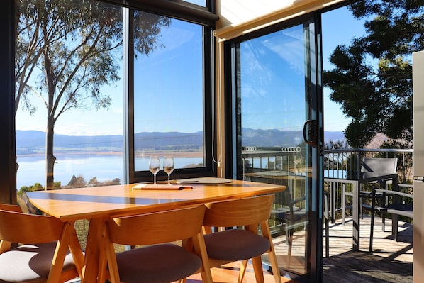A modern dining area with a wooden table and chairs, large windows with a view of a lake and mountains, and a deck with outdoor seating.