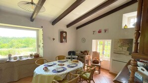 Zona pranzo con tavolo rotondo apparecchiato per quattro, circondato da sedie in vimini. Una finestra offre vista su piante verdi.