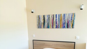 Un letto ben rifatto con una coperta grigia e due cuscini, un quadro astratto colorato sopra e una parete bianca con un interruttore.