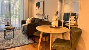 A modern living room with a sofa, a round table, and a small side table. There is a window with curtains, a wall-mounted air conditioner, and a framed artwork. - Lifestyle Apartment at Stationhouse (Cape Town)