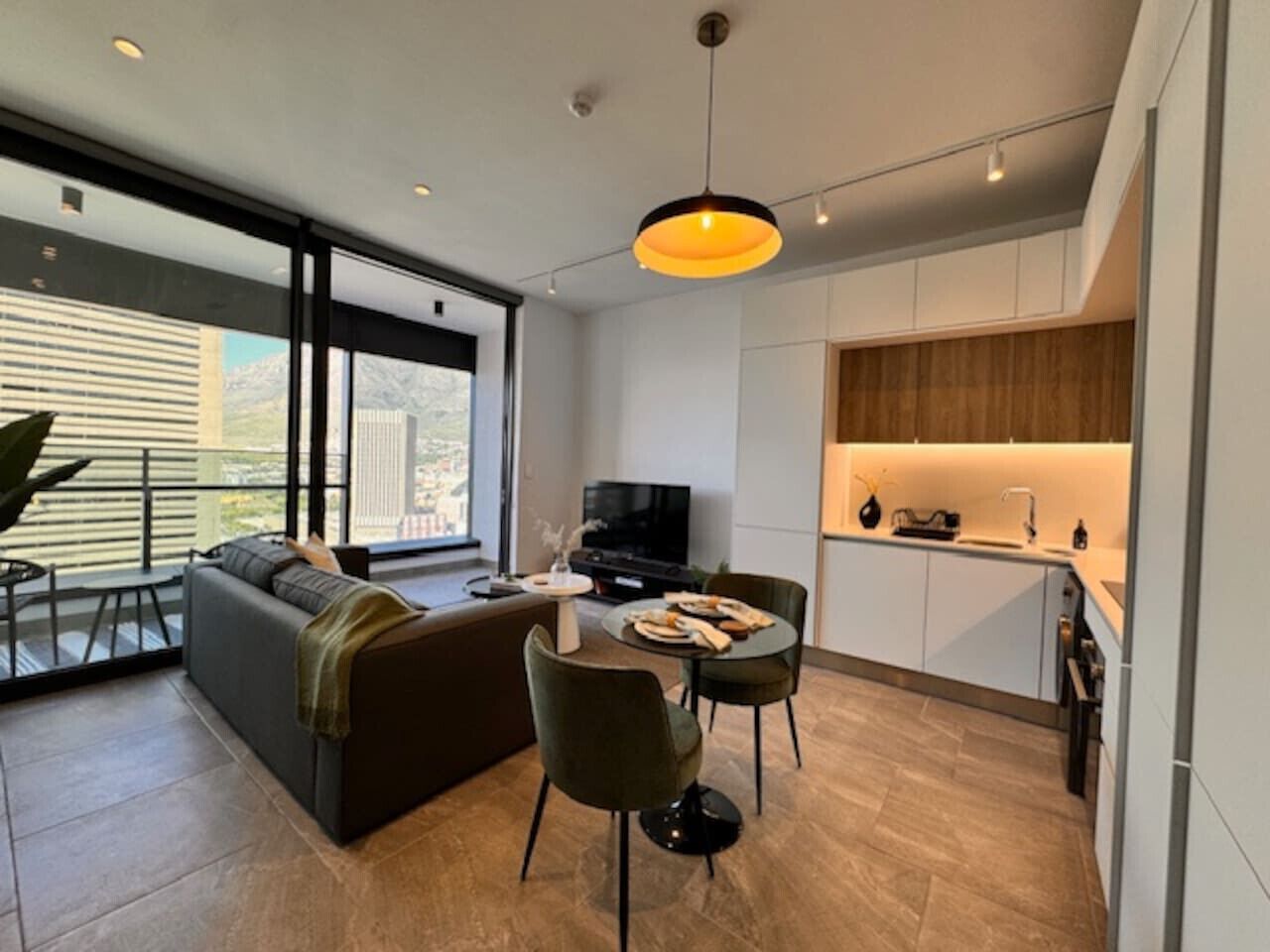 A modern living room with a sofa, dining table, and chairs, a TV, and a kitchen area with cabinets and a countertop.