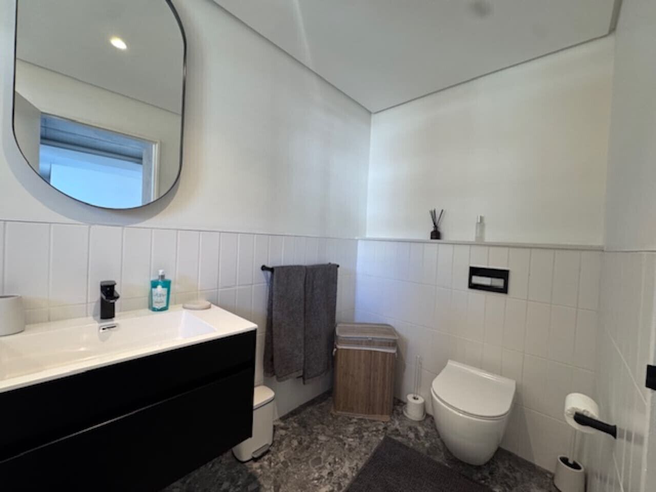 Een moderne badkamer met een witte wasbak, een zwart aanrecht, twee toiletten, een spiegel en een kleine prullenbak.