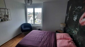 Un dormitorio con una cama, una silla, un ventanal y un cuadro.