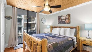 A bedroom with a wooden ceiling fan, a large bed with a quilt, a nightstand, and a view of a balcony with a railing.