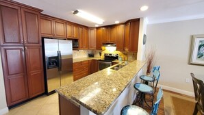 A kitchen with wooden cabinets, a stainless steel refrigerator, granite countertops, and a bar area with stools. - Ocean Reef 306 3 Bedroom Condo (St. Augustine)