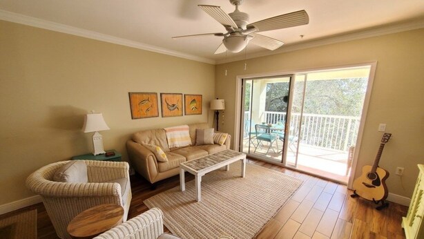 A living room with a beige sofa, a coffee table, a wicker chair, and a guitar. There is a ceiling fan, a window with a view of the outdoors, and a sliding glass door leading to a balcony. - Ocean Reef 306 3 Bedroom Condo (St. Augustine)