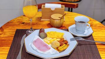Een ontbijt bord met eieren, ham, aardappelen en een stuk fruit, met een glas sinaasappelsap en een kop koffie op een houten tafel.