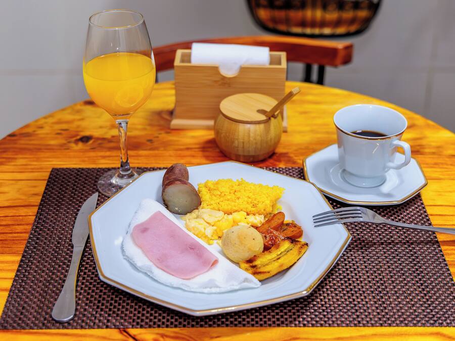 Een ontbijt bord met eieren, ham, aardappelen en een stuk fruit, met een glas sinaasappelsap en een kop koffie op een houten tafel.