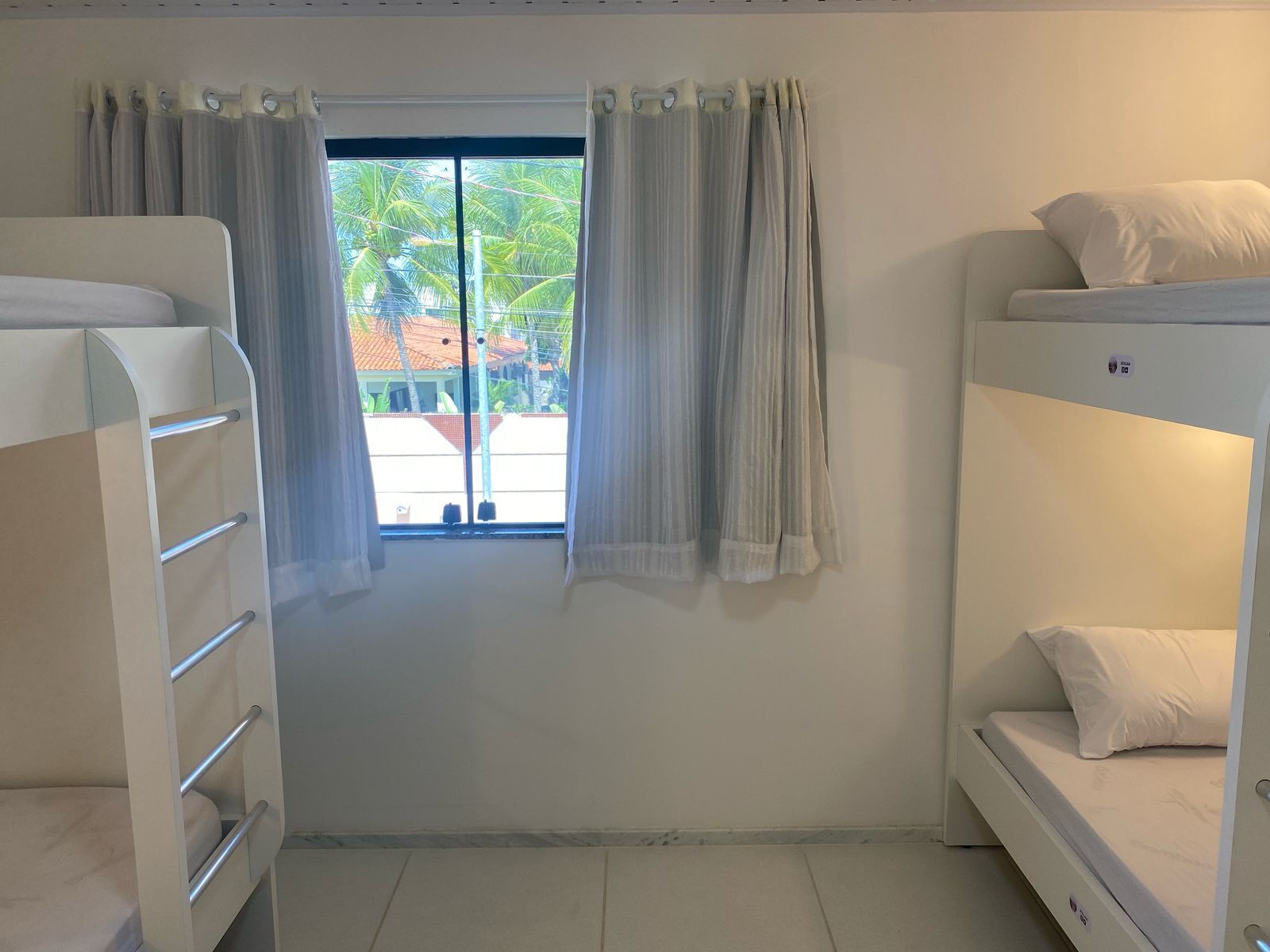 A room with bunk beds, a window with curtains, and a view of palm trees and buildings outside.