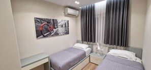 A small, modern room with two single beds, a bedside table with a lamp, a wall-mounted air conditioner, and a window with curtains. - Arkle Apartment 1 (Tirana)