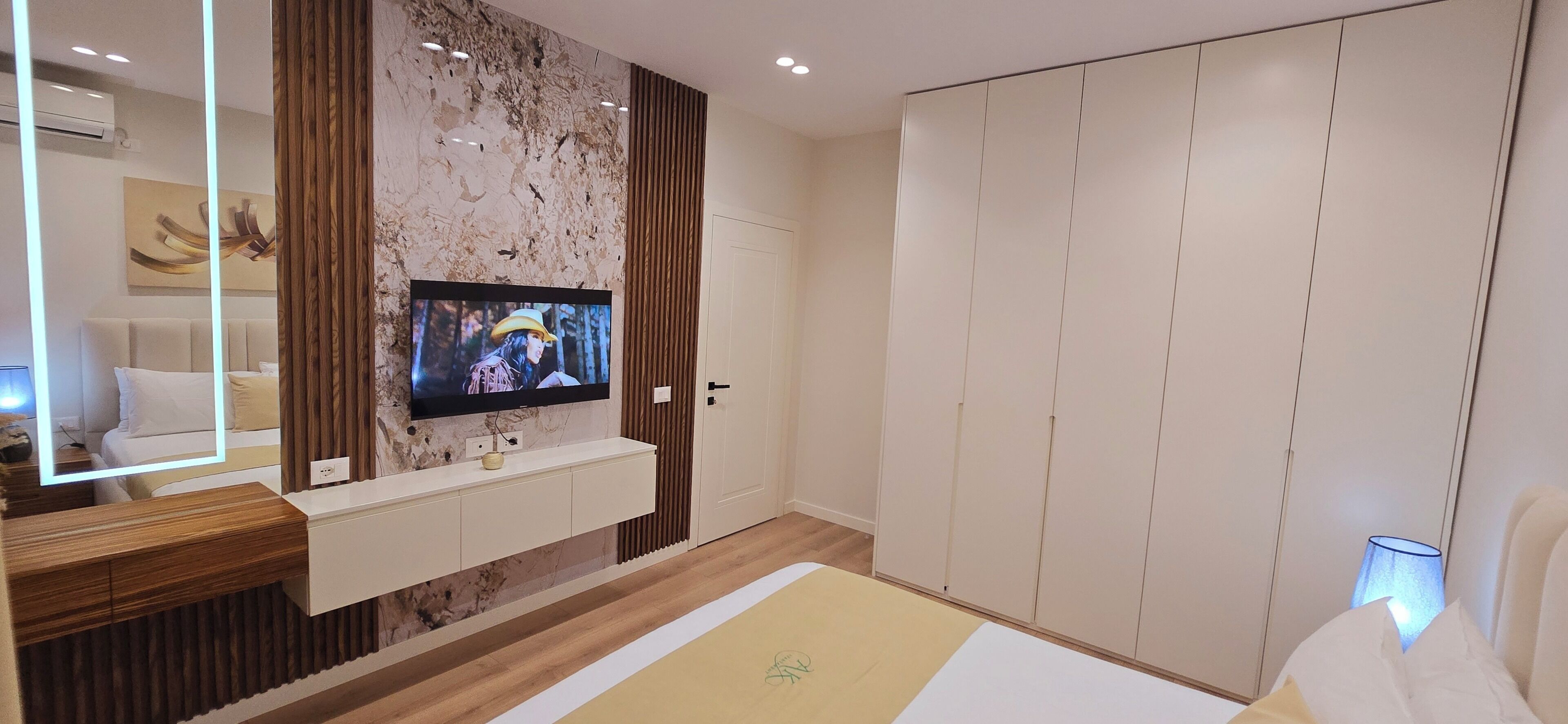 A modern bedroom with a TV mounted on a marble wall, a wooden vanity, a wardrobe, and a bedside table.
