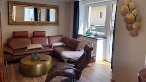 Ein Wohnzimmer mit einem braunen Ledersofa, einem runden Goldhocker, einem modernen Sessel, einem großen Spiegel und einem Fenster mit Blick ins Freie.