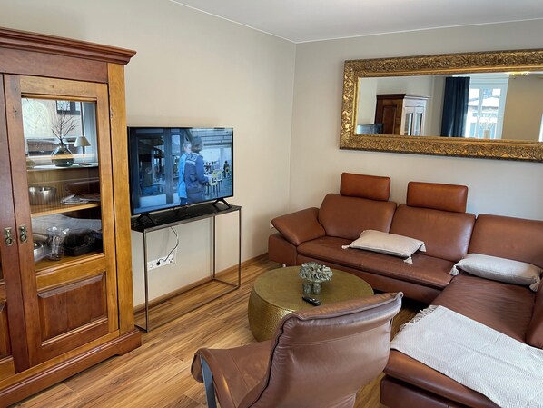 Un salon avec un canapé en cuir marron, un téléviseur posé sur un meuble, un buffet en bois aux portes vitrées et une petite table ronde sur laquelle se trouve un vase.