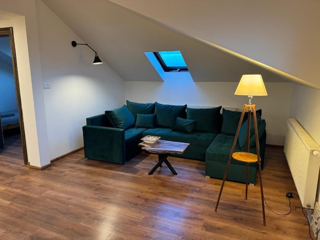 Un salon chaleureux, aménagé dans un style industriel, avec un canapé d’angle vert, une petite table basse, un lampadaire et une lucarne.