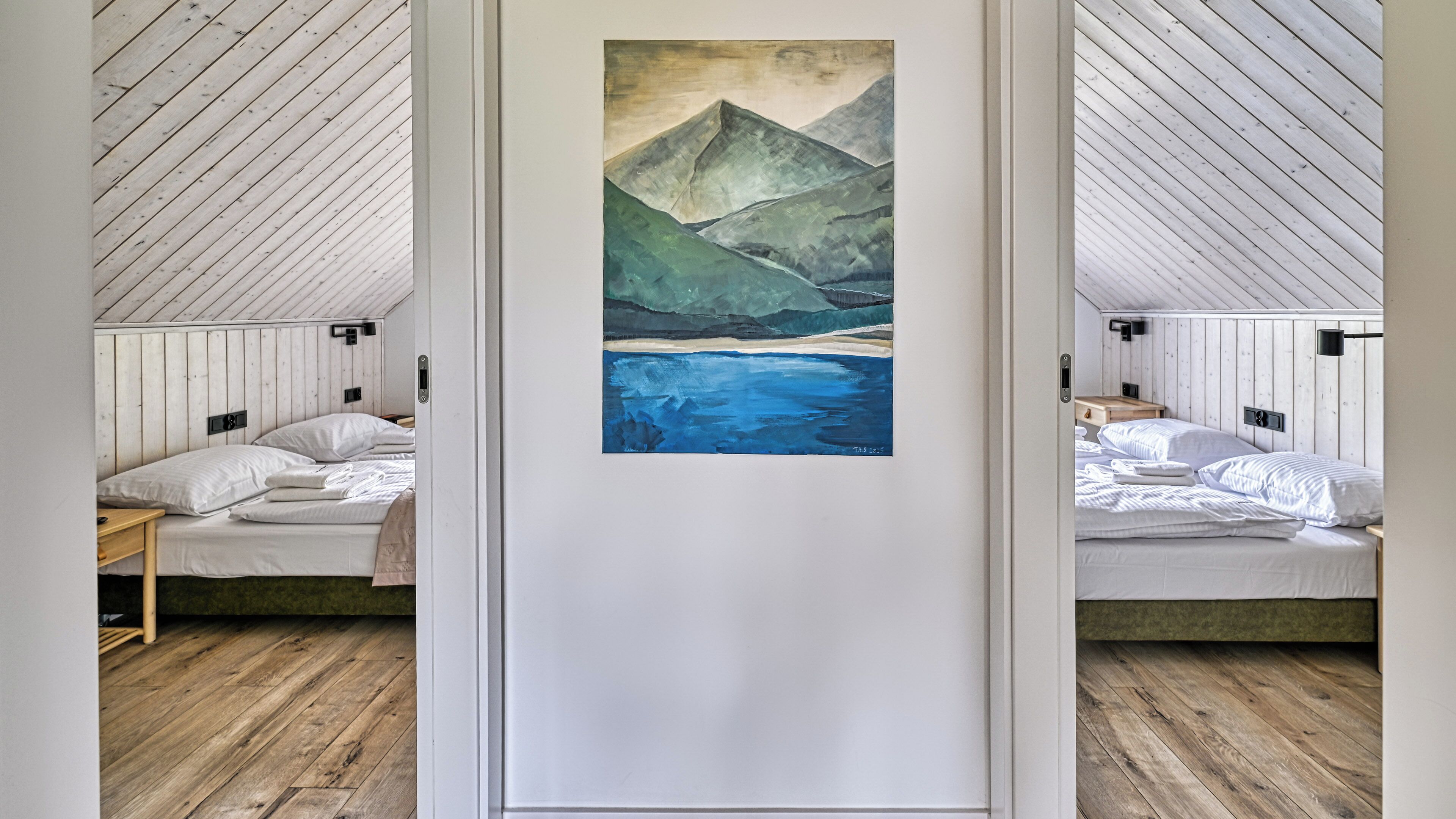 A room with a wooden sloped ceiling, a bed with white bedding, a painting of mountains and water on the wall, and a wooden floor.
