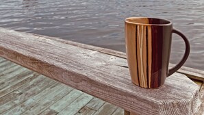 A wooden bench with a mug on it overlooking a calm body of water and distant hills. - Seaside Suites (Woody Point)