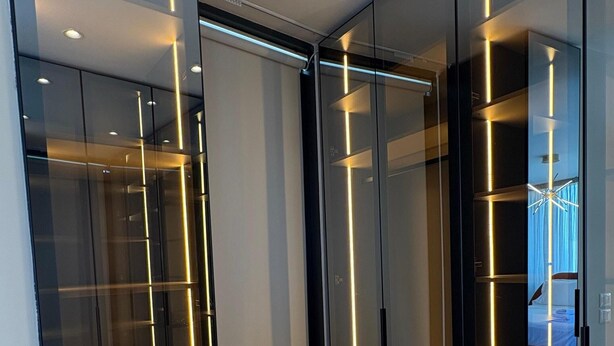Une chambre moderne avec un plancher en bois, une armoire en verre aux lignes illuminées et un lit agrémenté d’un rideau bleu.