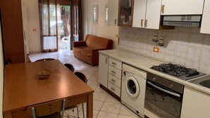A compact kitchen with a washing machine, a stove, and a dining area with a table and chairs. - Renovated apartment on the ground floor (Rosolina Mare)