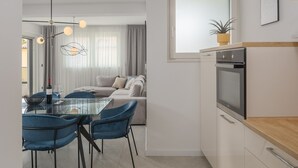 A modern kitchen with a built-in oven, a dining area with a glass table and blue chairs, and a living area with a sofa and curtains. - Apartman Blue in Poreč (Poreč)