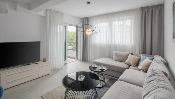 A modern living room with a flat-screen TV, a black coffee table, a beige sofa, and a large window with sheer curtains. - Apartman Blue in Poreč (Poreč)