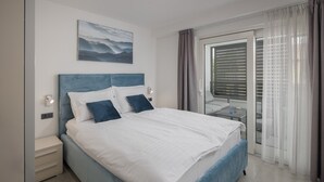 A modern bedroom with a large bed, a nightstand, a bedside table, a window with curtains, and a view of the outdoors. - Apartman Blue in Poreč (Poreč)