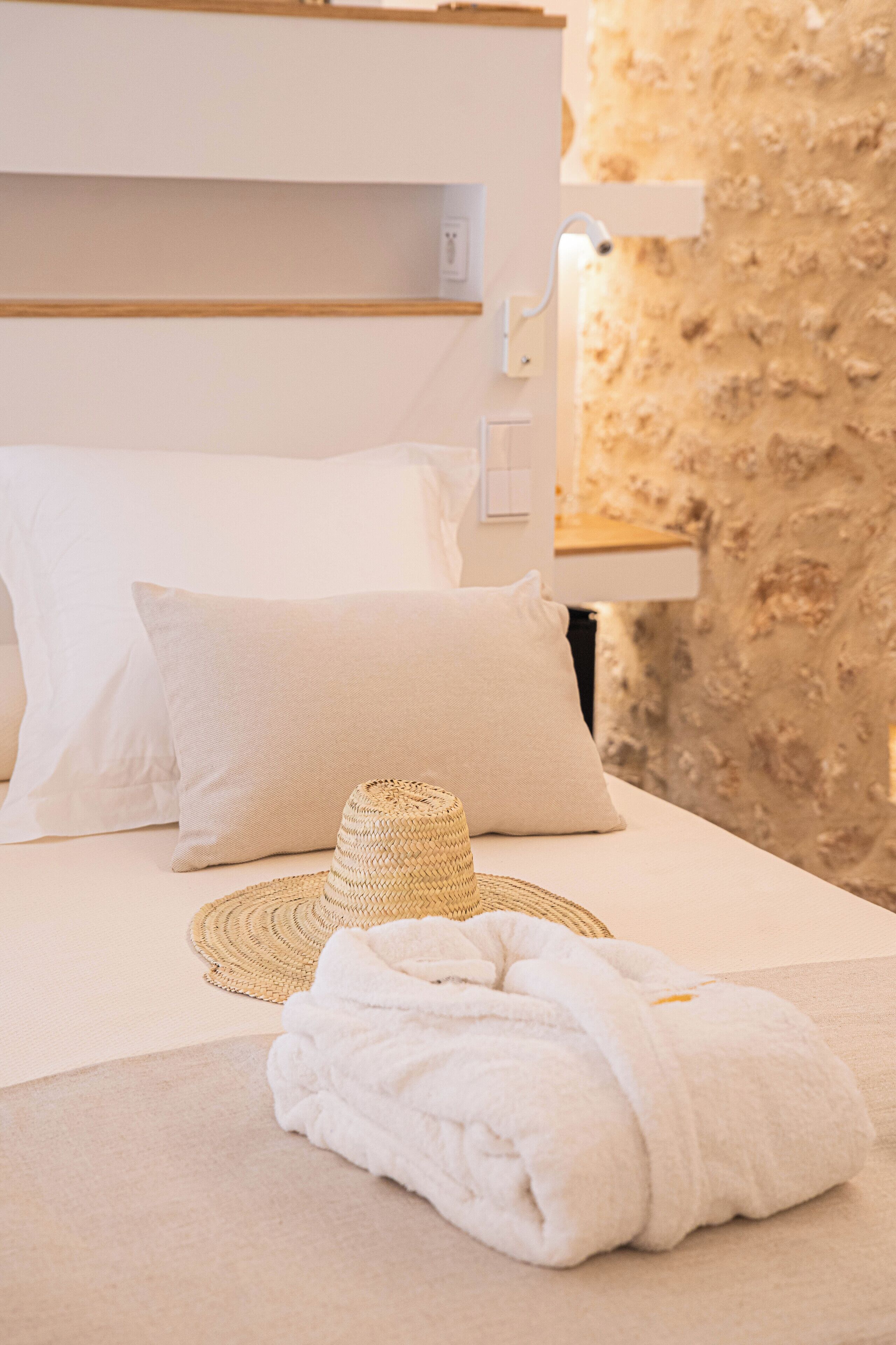 A neatly made bed with white linens, a woven hat, and a rolled towel.