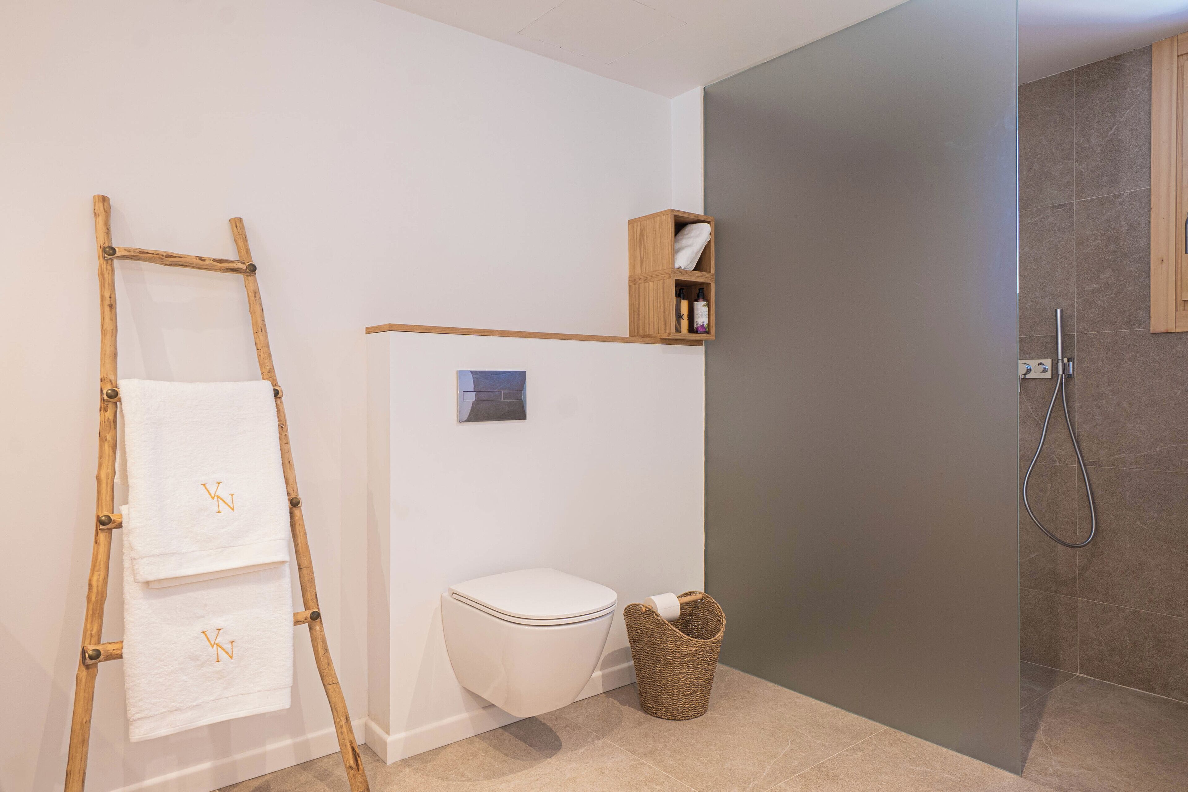 Un baño con inodoro, estante de madera con artículos de aseo, cesto de mimbre y zona de ducha.