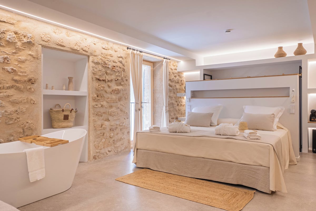 Une chambre moderne avec un mur en pierre, une baignoire, un lit avec du linge de maison blanc et une étagère ornée d’objets décoratifs.