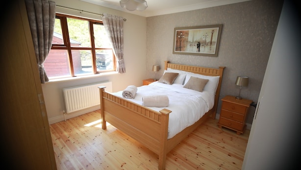 Een slaapkamer met een houten bed, een raam met gordijnen, een radiator, een nachtkastje, een dressoir en een schilderij aan de muur.